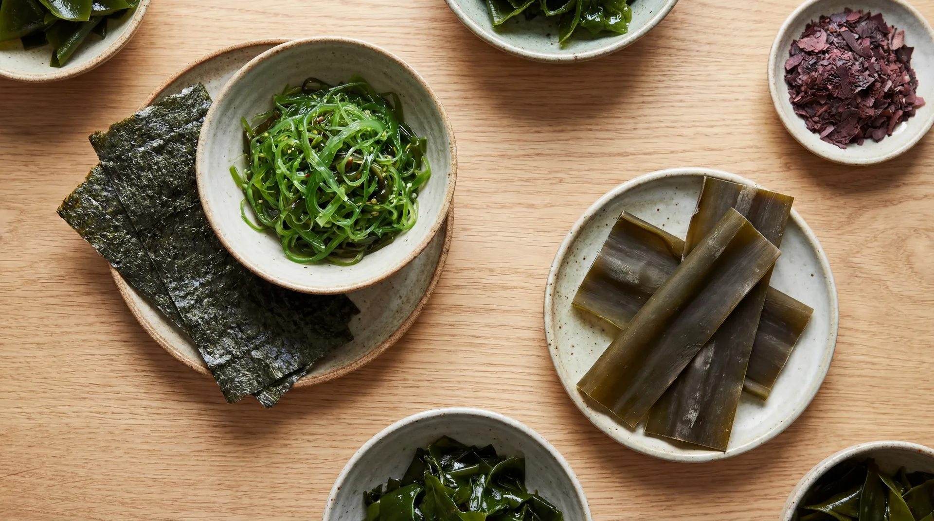 Assorted edible seaweeds in ceramic bowls on a wooden table, showing nori, wakame, kombu, and dulse.
